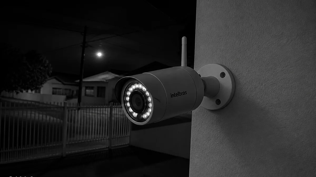 Câmera bullet externa com LEDs infravermelho acesos iluminando o muro de casa brasileira à noite, imagem em preto e branco no monitor de segurança.